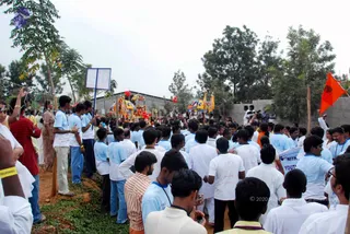 Outdoor photograph of a dense crowd moving along a dirt path between trees and a low wall during a procession. Many young men wear white shirts and light blue collared shirts with lanyards, some holding a blue-and-white banner reading YOUTH. In the centre, a decorated ratha or palanquin with colourful canopies, red and yellow drapery, and a trident-topped structure is carried forward. A saffron flag flies at right, and a few women in saris stand at the left edge. Given the crowd, the count is well above twenty.
