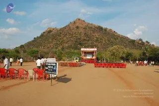 A photograph of an open dusty ground at the foot of a brown, scrub-covered hill under a partly cloudy blue sky. Rows of red and cream plastic chairs are set out on the left and right, facing a small ornate stage with a red-draped base and a gold-trimmed canopy, with a backdrop in the centre. A white sign with a red border and Tamil text points the way to the 1008 Ananda Lingam. A few people in white shirts and dhotis, and some women in saris, walk across the ground. Tall lamp posts line the path.