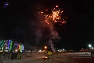Night photograph of an open dusty ground at an ashram or event site, with a burst of orange and red fireworks rising into a dark sky above a column of smoke. A yellow tractor sits at left in front of a multi-storey building covered in green, blue and red string lights. A small flame and smoke rise near a yellow structure at centre, and a few distant figures stand near vehicles at the right edge. Floodlights glow on the right. No one is clearly the central subject, and no identifiable person is clearly visible.