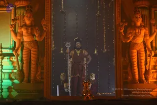 A standing figure stands centrally in a lit, dark-backed doorway framed by two large ornamental guardian statues with raised arms, one holding a mace. The central figure holds a silver trident, wears a crown-like headpiece with a crescent, a gold necklace, a rudraksha-style mala and a rust-red dhoti-style wrap, with a shawl over one shoulder. Hanging bells and strands of small lights decorate the backdrop, and brass oil lamps and a lion-shaped ornament sit at the base.