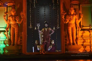 A figure stands at the centre of an illuminated shrine niche, holding a tall trident in the right hand and resting the left hand on the hip. He wears a maroon-brown dhoti, a maroon shawl draped over the shoulders, a crown, and multiple red and bead malas with a large gold necklace. Two large orange-lit stone dvarapala guardian statues flank the niche, each with four arms holding a mace, a trident-like weapon and other implements, wearing ornate gold jewellery. Strings of small lights and hanging bells decorate the dark backdrop, and a brass oil lamp stands at the lower right. The figure's appearance is consistent with a person but is uncertain, and no visible text is present.