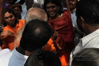 A crowded outdoor photo of a smiling young man with long dark swept-back hair, a red and gold brocade shawl over orange clothing, a gold necklace and a garland, with a red bindi. Beside him, a man in orange robes with long dark hair and a red tilak touches an elderly white-haired man in orange whose head is bowed. Other men in white shirts and a man in a plaid shirt press in around them, with a wooden pole visible at upper right. The surrounding people are watching and crowding closely.