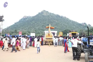 Outdoor festival scene at a dusty open ground before a large hill covered with trees. A crowd of many dozens of people in white dhotis and shirts, saris, and casual clothing mills about. In the centre, an ornate golden ratha (chariot) with a red-and-gold canopy and decorated gold figures stands, with a second decorated float behind it. A blue tractor with a trailer stands at right. Banners and a stage with lighting rigs frame the grounds, with a small portrait banner visible at left.