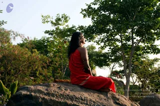 Outdoor photograph of a single person seated cross-legged on a large grey granite boulder amid green trees and shrubs at golden hour. The person has long dark hair falling loose down the back, wears a red draped garment and gold bangles on the arm, and gazes upward toward the sky, away from the camera. A small yellow-orange cushion or cloth lies beside the rock. A white bird-shaped logo appears at the upper left and a copyright line is overlaid at the lower right.