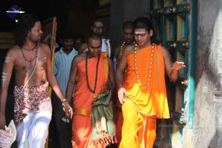 Photograph of a group walking out through a carved green-and-brown wooden temple door. In the foreground, a long-haired man in a rudraksha mala and ceremonial gold-bead necklace wears a saffron-orange dhoti and shawl with a white-and-gold cloth, holding his hand out in a gesture. Beside him, a shaved-headed man in orange robes and a rudraksha necklace walks with a green-bordered shawl. A bare-chested man on the left wears rudraksha beads and a patterned dhoti, with white marks on his arms. Several more men in orange and white follow behind, one with white forehead marks. Swamiji (probable) is the long-haired figure in orange with the gold-bead necklace.
