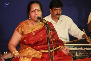 A woman seated on stage sings into a microphone on a stand, wearing a red silk sari with gold zari border and gold embroidered motifs, gold bangles, earrings and a necklace, with a red bindi and white forehead marking. Behind her, a man in a white shirt plays a percussion instrument, and a Roland keyboard is visible at right against a deep blue backdrop. The scene looks like a devotional musical performance.