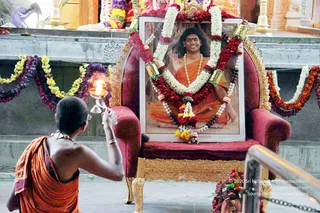 A devotee in a saffron/orange cloth, bare-shouldered and seen from behind, kneels on a grey stone floor and waves a lit oil lamp toward a large framed portrait on a red velvet throne with gold trim. The portrait shows a smiling figure with long dark hair in orange robes and a rudraksha mala, seated cross-legged, one hand raised in blessing. The portrait and throne are garlanded with white, red, yellow and purple flower garlands, and small brass lamps stand nearby. Behind the throne a wall of carved orange and gold temple decoration is visible. This is a puja offering to a portrait on a throne, a person appears as a depicted portrait, not a person physically present.