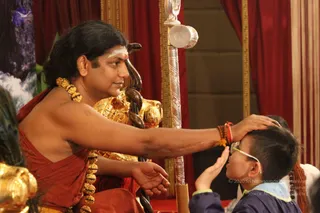 A seated man in an orange-red shawl and heavy gold necklaces places his right hand on the forehead of a young boy in glasses and a navy-blue top. The seated figure has long swept-back dark hair and a white-and-red tilak on his forehead, and a dark snake is coiled over the gold throne armrest beside him. A silver staff with a round finial stands behind the boy, red curtains hang in the background, and a peacock-feather fan appears at the left edge. Several other people are partially visible at the right. a person is blessing the child in what appears to be a darshan-style blessing.