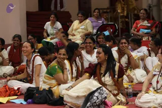 A photograph of a seated group of women at a satsang session on red mats in a hall with a wooden stage and gold-coloured altar elements behind them. Many women wear cream and gold-bordered saris or white outfits, some with rust and maroon shawls, and several have name badges clipped to their clothing. Most are smiling or laughing, one woman in a green and gold sari covers her mouth, and a water bottle sits on the mat. A woman in a red saree sits in the background on a red chair. Several women in the front row are laughing together in a relaxed, joyful atmosphere.