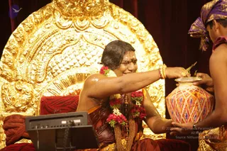 A photograph of a seated figure on an ornate gold throne with a red cushion, dipping his hand into a decorated pot with pink, orange and blue geometric patterns held by a young attendant. The seated figure wears a maroon and orange shawl, multiple red and white flower garlands, a gold necklace and gold bangles, and has a wet head with a white forehead mark. The attendant wears a purple and gold head wrap and is bare-chested. Behind is a red curtain and a black monitor. The scene suggests an abhishekam-style ritual with water from a kalasha.