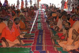 A large seated gathering of sadhus in saffron and orange robes sits in rows on colourful striped woven mats during a satsang-style session. Several men in front wear orange garments and a few wear white dhotis, with tilak marks on foreheads and rudraksha malas visible. Some hold open books or papers. Along the centre, a long striped cloth runs down the aisle between the rows, with brass plates placed on the mats. Behind them, standing figures in white and orange stand under a tent canopy with red chairs and a red-orange cloth hanging at the back.