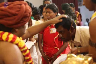 A photograph of a blessing line at an indoor event. In the foreground, a man in an orange-red turban and a yellow-red marigold garland stretches his arm to place his hand on the head of a kneeling man in a white shirt and dhoti with a rudraksha-style cord. Behind them, a woman in a red sari with a gold border and a visible name badge stands with her hands clasped, while a woman in white with a white shawl holds a paper and a bead mala waits in line. Other devotees in saris stand in the background against a maroon curtain. A gold-coloured vessel is visible at the lower right.