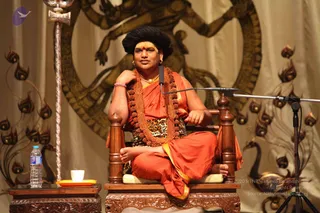 A seated figure in an orange saffron robe and rudraksha mala sits in a carved wooden armchair on a stage. He wears a leopard-print cloth at the waist, a gold necklace, and a yellow tilak with a red mark on the forehead, with his hair tall and dark. A microphone stand is at right and a water bottle and small white cup on a tray sit at left beside a silver staff. The backdrop shows a golden Nataraja-style dancing figure and stylised peacocks. a person cues: ornate saffron clothing, rudraksha malas and gold necklace, shown as a seated speaker on stage.
