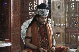 A man seated in meditative posture with eyes closed, wearing a leopard-print headwrap, an orange-saffron shawl over bare shoulders, and multiple tiers of rudraksha beads and gold-tone beaded necklaces. Rudraksha bands are worn on his upper arms and wrists. A silver-rimmed carved wooden chair and an ornately carved wooden screen stand behind him, with a microphone on a stand to his right and a small red cup on a silver tray at left. Probable Swamiji by appearance cues: long swept-back hair, saffron clothing, rudraksha malas and ceremonial gold necklaces, seated in a ritual setting.