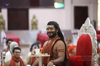 Indoor satsang hall with a seated man in the centre on an ornate silver-framed throne with red cushioning. He wears a saffron-orange draped robe, a rudraksha-style beaded necklace, a gold necklace, a gold armband and a white tilak with a red dot on his forehead, with long dark hair. A white cup sits on a silver side table beside him. Behind him, devotees in orange and red shawls and white vibhuti marks sit on red-covered mats, with a golden deity murti visible in the background.