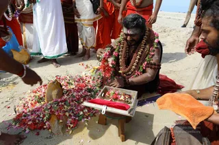 Outdoor photo on a sandy beach of a man kneeling before a wooden-framed tray holding flowers and a red cloth, beside a large coconut and heaped red, pink and white flower petals. He wears a long mala of rudraksha beads with gold-coloured ornamental beads, a garland of pink and white flowers, a tilak mark on his forehead, and orange-red cloth. Around him, several people in orange robes, white dhotis and gold-bordered clothing stand barefoot on the sand. An orange towel is held at the lower right. The scene resembles a beach puja or abhishekam offering. A person in the foreground at the lower right wears a red cloth and beads.