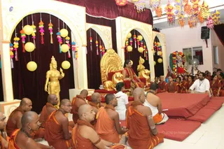 A satsang in an ornate hall with a golden throne backdrop. A seated figure with long dark hair wears a red and orange garment with ornate gold and beaded necklaces, seated on a gold throne with red cushions, flanked by golden lion-form and golden deity statues. In front, a large group of shaven-headed men in saffron-orange robes with rudraksha malas sit on the floor, and devotees in red and orange attire sit along the right. A decorated deity murti stands at the right edge. Hanging yellow paper lanterns, tassels and colourful paper ornaments decorate the arches and ceiling. A few white-clad attendants sit near red floor cushions. The central figure's appearance is consistent with the page's identification but uncertain from the image alone.