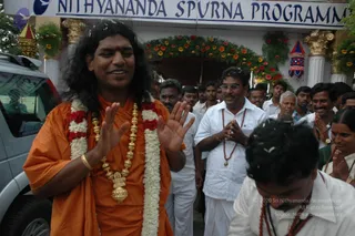 A man with long, swept-back dark hair stands at left in saffron-orange robes, a white-and-red flower garland over his shoulder and a heavy gold ceremonial necklace with a large pendant at his chest, hands raised in greeting near his face. Behind him, a crowd in white shirts and dhotis, some wearing rudraksha malas, walks past a flower-decorated gateway beneath a banner reading Nithyananda Spurna Programme. Flower-garlanded columns, a small lantern-shaped decoration, and a silver van at left complete the scene. A man in front right bows with folded hands, and a woman in a white sari with a rudraksha mala stands at right with palms joined.