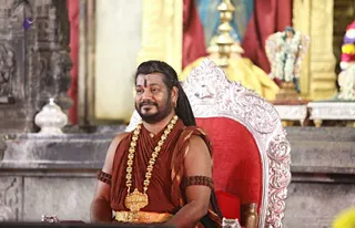 A man seated on an ornate silver throne with a red velvet cushion inside a temple hall. He wears a rust-brown and orange shawl draped over his shoulders and torso, a long gold beaded necklace with a large gold pendant, gold bangles and rudraksha-style armbands, and a dark forehead mark. His long dark hair is swept back, and he has a full beard. Behind him stand gold pillars, red drapes and a flower garland, and to his right is a decorated shrine area. The figure matches Swamiji (probable) by his long swept-back hair, ornate gold necklace and saffron-orange clothing, seated on a throne in a temple setting.