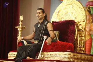 A seated man sits on an ornate gold throne with a deep red velvet cushion and red-and-black zebra-patterned upholstery, speaking at a satsang stage. He wears a sleeveless black kurta with silver embroidery, a black-and-gold waistband, a rudraksha mala and a silver pendant, and a red bindi on his forehead. He has long hair tied back and a small headset microphone, and holds a silver staff across his lap. A brass oil lamp stands at left beside red curtains, and a painted deity figure with a tall crown appears at right. Swamiji (probable): the long hair, rudraksha mala and ceremonial ornaments on the throne suggest the teacher of the satsang, seated in a position of honour.
