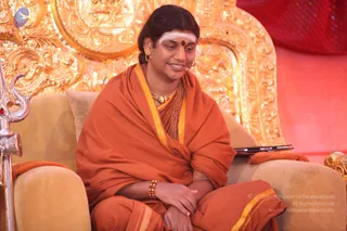 A seated man in an orange saffron shawl with yellow borders sits on a cushioned golden armchair in front of an ornate gilded throne backdrop with carved figures. He wears a white forehead marking with a red bindi, gold hoop earrings, a gold and beaded necklace, and a rudraksha mala-style bead bracelet. His hands rest in his lap and he smiles while looking to the side. A silver trident-topped staff stands at the left edge, and a red curtain fills the right background. Swamiji (probable) in_person seated on the throne, identified by long swept-back hair, saffron clothing, rudraksha beads and ceremonial gold necklace.