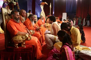 A photograph of a ceremonial gathering on a red-carpeted stage with red drapes behind. Several men in saffron-orange robes sit on ornate gold-and-red thrones; the foreground figure on the left wears orange with a gold elephant-shaped armrest. Behind them a white-robed man with dark curly hair kneels, and a man in a yellow dhoti-style garment bows forward at the lower right. A woman in a pink sari with gold border kneels in the foreground, and another woman in a green sari stands at right near a camera tripod. Gold ornate staffs and a golden cobra-hood canopy stand behind the seated figures. Some seated men wear dark-framed glasses and have grey beards. The scene suggests a seated ritual reception with devotees paying respect.
