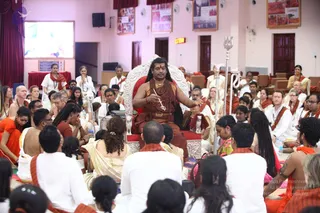 A seated man in a white-and-silver ornate throne (peetha) addresses a large seated audience in a hall with pink walls and red curtains. He wears a saffron-orange-red garment, a red-and-white tilak on his forehead, layered rudraksha malas and a gold necklace, and holds a small ceremonial stick. A tall trident-topped staff stands beside him. Around him, devotees in white clothes with saffron-orange shawls sit on the floor, and some hold water bottles. A wall-mounted screen shows an image, and a banner on the wall reads Sannyas. A clock displays 11:59. A long-haired figure with swept-back hair and ornate attire on the throne matches the Swamiji (probable) cues of saffron clothing, rudraksha malas and gold ornaments, presiding as a discourse speaker.