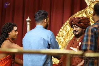 Photograph of a stage event with a red draped curtain and an ornate gold throne-like chair. A seated man in a brown-maroon turban and rust-brown robe, with a gold chain, smiles with eyes closed while a man in a light blue shirt stands in front of him. At left, a man with long dreadlocks, white tripundra marks on his forehead, a rudraksha mala and an orange cloth draped over his body reaches toward the scene. At far right, a man in a plaid shirt is partly visible. A brass rail runs across the foreground.