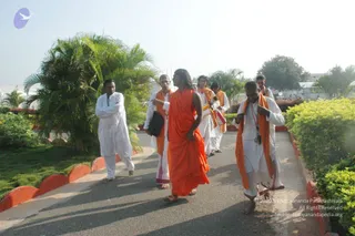 Outdoor photo of a group walking along a paved garden path lined with green shrubs, palm trees and red-edged kerbs under a hazy sky. In the centre, a man with long swept-back hair wears a flowing saffron-orange robe draped over his body and walks barefoot; the appearance cues are a long hairstyle and saffron clothing, so he is a person figure, though the identity is uncertain. Behind him, several men in white dhotis and orange shawls follow, one carrying a black bag, and a man in white with a shawl stands at the left edge.