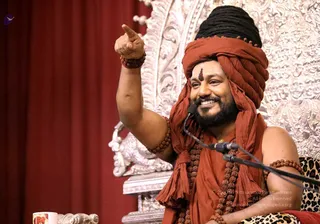 A smiling bearded man in a rust-orange turban and matching orange shawl sits on a leopard-print seat before an ornate silver throne with repoussé floral carving and a red curtain behind him. He raises his right arm and points upward with his index finger, wearing rudraksha beads on his wrists and a long rudraksha mala across his chest. A microphone is positioned near his mouth, and a small cup sits at the lower left. Probable Swamiji (appearance cues: orange turban and shawl, rudraksha malas, speaking from a silver throne), seated and giving a satsang talk. The image is a photograph of a single speaker.