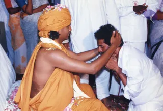 A seated man in a saffron-orange turban and saffron-orange wrap, bare-chested beneath it, with a white flower garland around his neck and petals on the ground, places both hands on the head of a kneeling man in a white shirt and white dhoti. Behind them, several people in white and cream clothing stand or kneel in a crowded hall. The scene appears to be a blessing or energy darshan moment with flower petals scattered around.