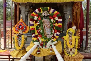A deity murti stands centrally in a shrine heavily decorated with a large arch of marigolds, red roses, white flowers and green leaves, with a kalasha-like crown and gold ornaments. Two smaller attendant murtis flank it, each wearing yellow and green marigold garlands, one holding a red and blue canopy. Yellow cloth drapes the central figure and white ritual ropes lead toward it. Behind are stone pillars, a brass lamp stand and a red umbrella. Visible signage reads Nithyananda Dhyanapeetam and Ayurveda-related text. No people are visible; figures are statues.