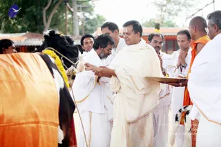 A photograph of a ritual at an outdoor ashram setting with a black-and-white cow draped in an orange cloth and garlanded with yellow flowers. A man in an off-white dhoti and shawl, with a tilak on his forehead, holds a brass plate and offers to the cow while another man in white stands beside him. Several men in white dhotis and shirts stand around, some with hands folded, and one man in orange clothing stands at the right. Red-roofed buildings are visible in the background.