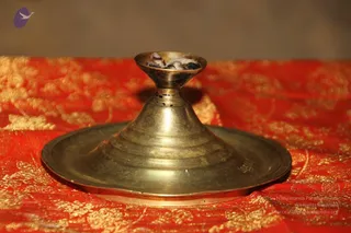 A close photograph of a brass or bronze oil lamp with a wide circular base and a tall conical stem, holding a small cup of burnt wick residue. It rests on a red fabric with gold floral brocade pattern. No people are visible, and the background is blurred dark grey.