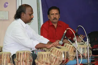 Two men sit side by side playing tabla drums in front of microphones on stands, in a performance setting. The man on the left wears a white long-sleeved shirt and bends forward over the drums, hands pressed on the drumheads. The man on the right wears a red shirt with a small red bindi on his forehead and plays the drum at right while looking toward the camera. A blue backdrop hangs behind them, and a black bag with a red logo sits at the right edge.