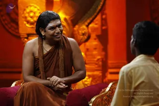 A seated man in a rust-brown saffron-toned robe draped over one shoulder, wearing a long rudraksha-style mala of brown beads and a thin necklace, with a red-brown tilak on his forehead, leans forward and smiles while speaking. His hands are clasped in his lap. He sits on a gold-trimmed throne-like chair with deep red cushions, in front of an ornate orange-gold carved backdrop. Facing him from the right, partly visible, is a younger person in a pale cream shirt. The setting resembles a discourse or satsang stage.