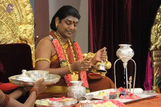 A seated man in a rust-orange and gold dhoti-style cloth, wearing a red-and-white flower garland, a gold chain with rudraksha-style beads, gold armbands and bangles, and a red bindi, holds a silver handbell and small silver vessel over a tray of flowers and offerings during a puja. A gold-carved throne with lion motifs and red velvet curtains frame the scene. In the foreground, a hand holds a silver tray with a covered silver bowl. A silver kalasha on a stand and a small figure stand on the ritual tray beside marigold and pink petals.