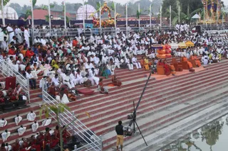 Outdoor photo of a large ceremonial gathering at a stepped red-stone tank or pool at an ashram campus. Hundreds of people in mostly white clothing and some orange, red and green saris sit on tiered steps facing a raised platform at the top right. On the platform, several figures sit on ornate chairs under decorative orange and yellow parasols; one person in orange robes is seated near the front. A camera operator in a dark shirt and yellow shorts stands on the lower steps beside a tripod-mounted camera and a boom pole. Orange-robed monks sit at the bottom left, and white-clad helpers are visible along the steps. Flags, colourful arches and a festive temple-style structure decorate the background.