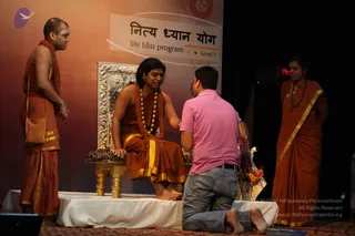 Stage event with a backdrop reading "नित्य ध्यान योग" and "life bliss program Level 1" beside an Om symbol. A long-haired figure in orange-saffron robes with gold borders sits on an ornate silver-and-gold throne with a gold-legged table, wearing a long rudraksha mala and a gold necklace. A man in a pink polo shirt and jeans kneels before the throne, reaching toward the seated figure. To the left stands a shaved-headed attendant in orange robes and a rudraksha mala, and to the right a standing attendant in orange sari-style robes with a gold border and beads. A flower arrangement sits at the right edge of the stage. a person seated as the guru, with cues of long swept hair, ornate gold necklace, rudraksha mala and saffron robes.