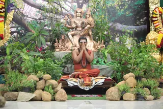 A seated man in saffron-orange robes with a rudraksha mala sits with palms pressed together in namaste on a tiger-skin-draped seat, a white vertical mark on his forehead. He is surrounded by potted plants, garden stones and flowers in a lush setting. Behind him stands a large golden murti of a deity pair, with smaller golden figures around its base, against a painted backdrop of a banyan-like tree. Garlands of yellow and red flowers hang at the edges.