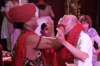 A photograph of a ritual exchange in a dim, reddish-lit hall. In the foreground, a woman wearing a red turban, a red shawl printed with script, a rudraksha bracelet and a leopard-print top holds her hands to the face of a bald, bearded man in a white shirt, who wears a red shawl with script and a red rudraksha bracelet. Behind them, several people in white and pink clothing stand and watch, and a woman with short reddish hair and a grey scarf looks on at right. Brass items and offerings sit at the lower left.