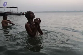 A man with wet, slicked-back hair stands waist-deep in a murky river at dawn, palms pressed together at chest height and face lifted upward in prayer, wearing a wet white cloth around his waist. Behind him, another bare-chested man wades in the water, and a covered wooden boat sits moored on the far bank under a hazy grey sky. The scene suggests a ritual bath in the Ganga, with no visible throne, saffron robes, malas or other markers identifying a person candidate.