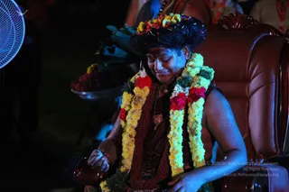A photograph of a smiling person seated in a large brown leather throne-style chair at night, with blue-lit surroundings and a fan at the left edge. The person wears a crown-like headdress decorated with flowers and sprigs, layered garlands of yellow marigolds with red roses and white blossoms, a rudraksha mala, a pendant, and a dark wrap with a patterned lower garment. A silver tray of flowers and leaves sits behind. Swamiji (probable) is described by the crown, flower garlands, rudraksha mala and ceremonial pendant, seated on a throne during a mandala process.