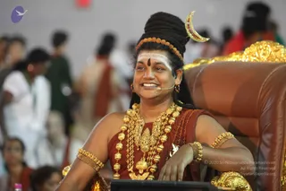 A smiling man with long dark hair piled up and a crescent moon ornament and a rudraksha headband sits in a brown leather chair with gold carved armrests. He wears ornate gold necklaces, gold bangles, a rust-red sleeveless garment and a small headset microphone, with white tilak markings on his forehead. Behind him, blurred people in white, green and red clothing stand in a crowded hall. a person: the seated man is shown in ceremonial attire with a crescent, rudraksha and gold ornaments, seated on a throne-like chair.
