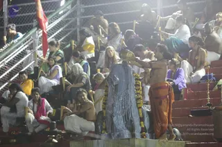 Night photograph of a large dark stone Shivalinga being bathed with water at an outdoor ritual, decorated with a marigold garland. A bare-chested man in an orange dhoti stands at right, holding a brass vessel raised above the linga, while a crowd of devotees in white and pastel clothes sits on red tiered steps behind a chain-link fence. Brass lamps stand along the steps, and a red flag hangs at left. Several people hold small brass lamps or vessels. The scene suggests an abhishekam performed by many participants.