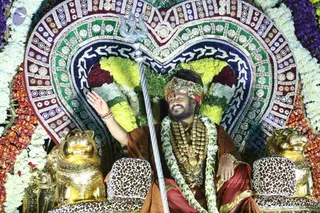 A seated man in an ornate procession palanquin, decorated with a heart-shaped embroidered canopy in red, green, navy and cream, framed by white and orange flower garlands. He wears a gold crown with a crescent, a trident (trishula) raised beside him, a heavy gold necklace and multi-strand rudraksha-style beads, and a rust-orange wrap over a red garment. He raises one hand in greeting. Two golden lion figures flank him, and the seats have leopard-print cushions.