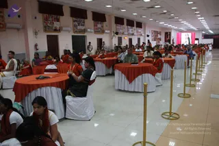 A large hall with tiled floor and rows of round tables draped in orange cloths with white skirts. Seated attendees, many wearing orange and saffron shawls, sit at the tables and watch toward a screen at the far end showing a figure in red. Brass stanchions with ropes line the right aisle. Framed photo panels hang on the walls and red drapes frame the stage area.