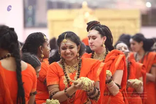 A joyful group of women in saffron-orange saris and blouses gathers in a hall during a Krishna Janmashtami celebration. The central woman smiles broadly, wearing gold jhumka earrings, a gold necklace, multiple rudraksha malas and gold bangles, and holds a handful of white-green offering. Beside her, a woman with a long braid and forehead tilak clutches the same kind of offering, and others in orange stand around with rudraksha malas and bindis. A large golden structure glows in the blurred background. A small logo appears at top left.