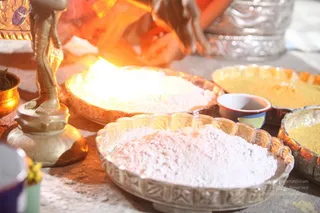 Close photo of an Aushada preparation process on a table. In the foreground, a large silver-rimmed bowl holds a heap of fine white powder, and beside it a shallow bowl holds yellow powder. A brass lamp stand with a figure is at the left edge, and a small brass vessel with green leaves sits beyond it. Blurred hands in orange sleeves reach over a bowl of white powder with a bright flame glow behind. No faces are visible. A small cup and additional metal bowls of powder complete the arrangement.