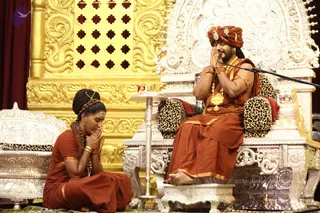 A photograph of a seated man in an ornate silver throne with leopard-print cushions, wearing a rust-orange turban with gold ornaments, orange robes, gold necklaces and bangles, with palms pressed together in greeting before a microphone. Beside him on the left, a woman in a rust-orange sari with a rudraksha mala and a tilak on her forehead kneels on the floor with her hands folded in namaste. Behind them stands a large gold-and-silver ornamental backdrop with carved lattice panels. A small table holds a framed picture. Swamiji (probable): the seated man wears a turban, orange robes and ornate gold necklaces, with a microphone set before him in the discourse setting.